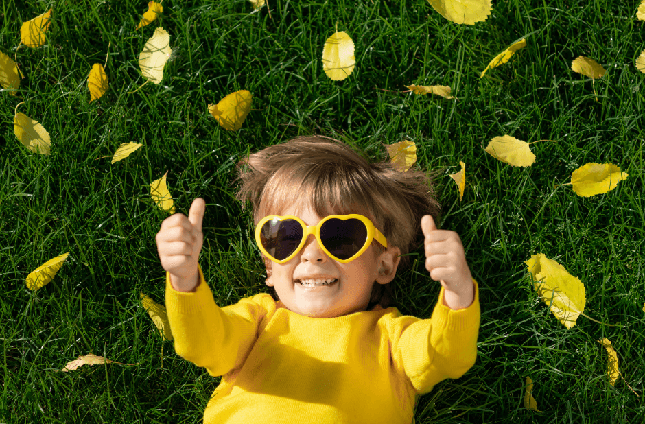 A kid in yellow lying in the grass in yellow leaves with shades with thumbs up and approves the affordable health insurance