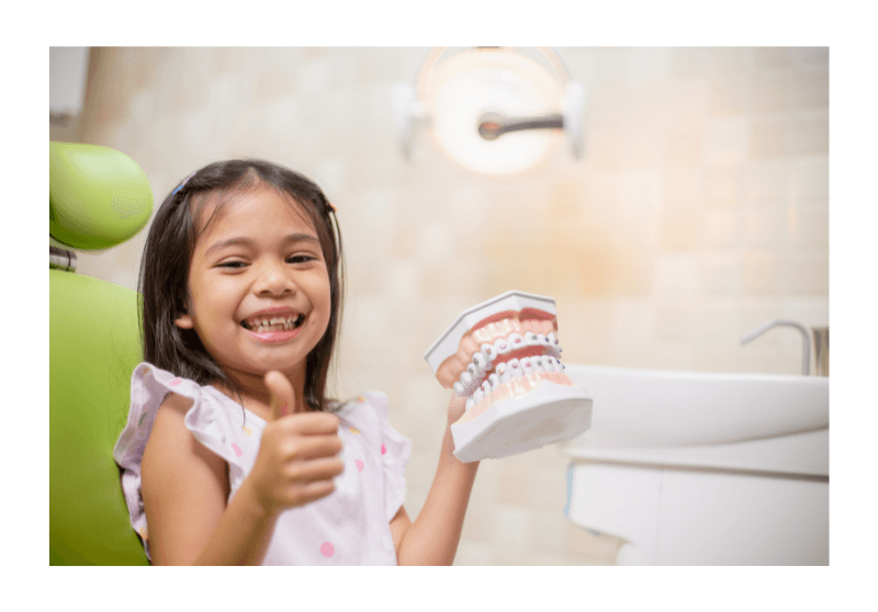 A girl with a thumbs up holding a teeth model in a dental clinic using her dental insurance plans to prevent tooth decay