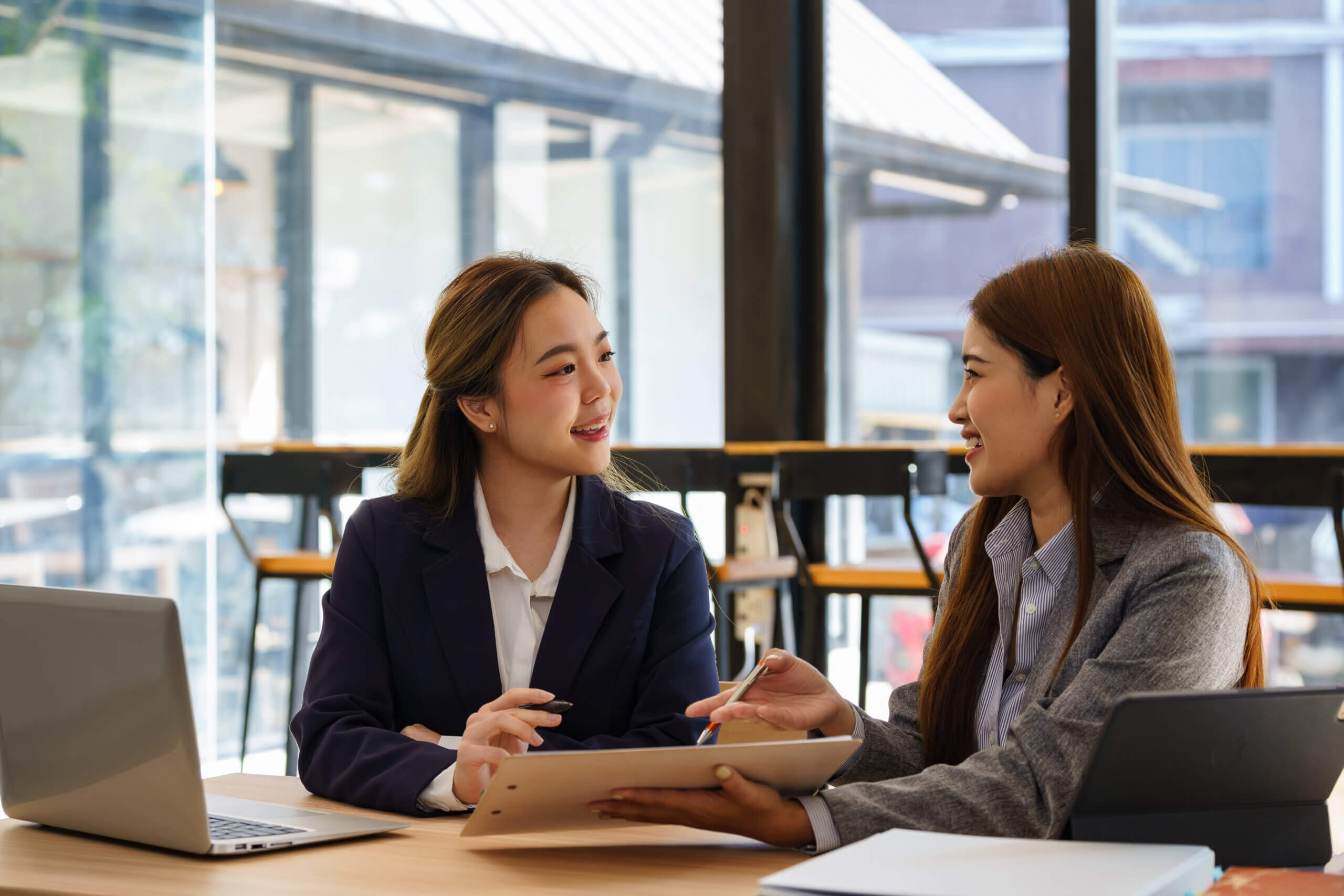 Two Asian businesswomen sit at desks discuss project details, share opinions, and work on collaborative tasks.