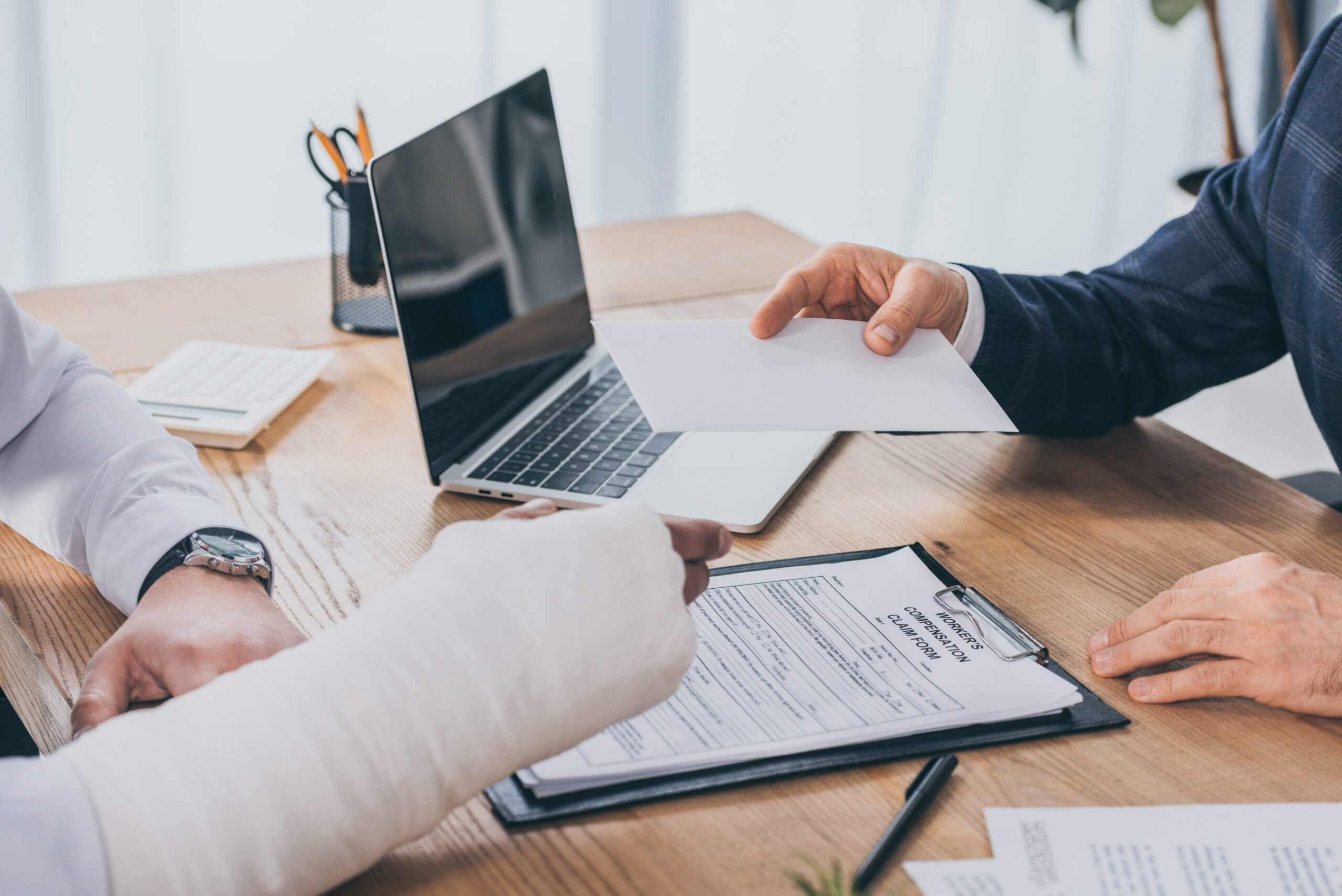 cropped view of businessman sitting at table and giving paper sheet to worker with broken arm in office, compensation concept covered by workers compensation